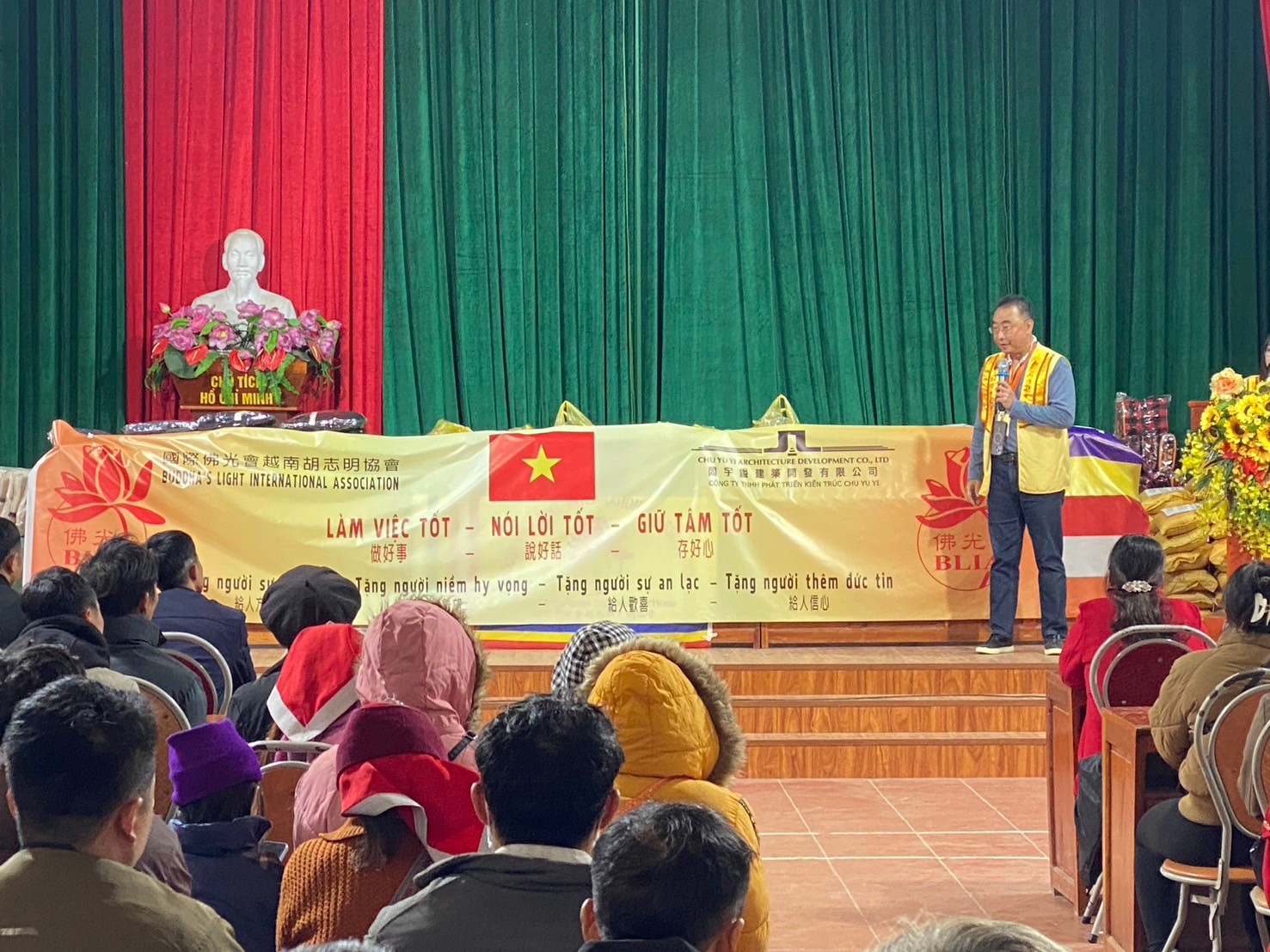 A person standing on a stage with a banner in front of a crowd

Description automatically generated